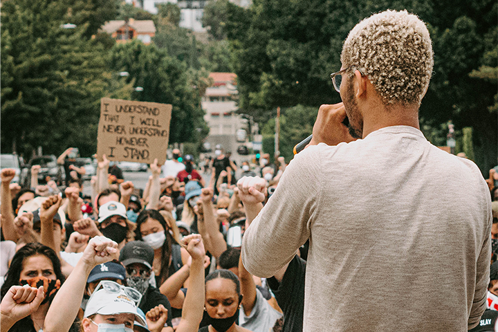 A man with blond curly hair speaks into a microphone facing a crowd at a protest. Many people raise their fists, and one holds a sign reading, “I understand that I will never understand. However, I stand.” Trees and buildings are visible in the background.