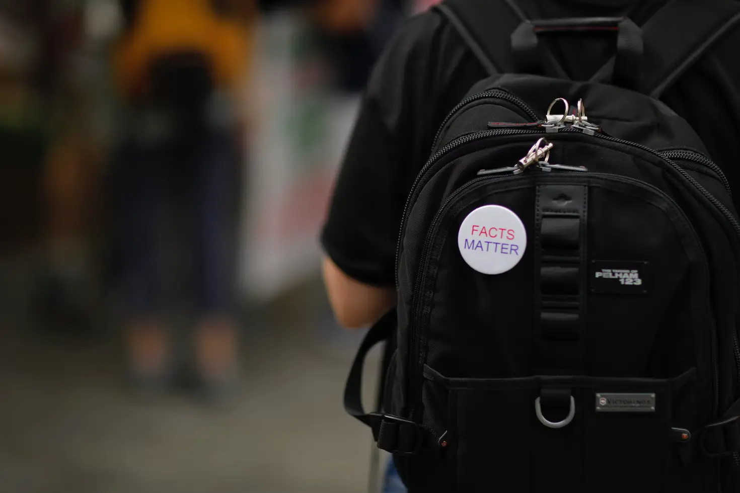 A person wearing a black backpack with a pin that reads FACTS MATTER. The background is blurred, focusing attention on the backpack and pin.