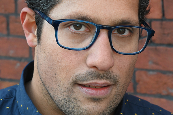 Close-up of a man wearing blue-rimmed glasses and a navy shirt with small yellow dots, standing in front of a red brick wall. He has short dark hair and is looking slightly to the side.