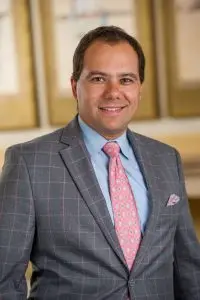 A man in a gray checkered suit with a light blue shirt and a pink patterned tie smiles at the camera. The background is softly blurred with gold and beige tones.