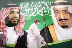 People wearing face masks walk past a large billboard featuring two Saudi leaders in traditional attire, set against a green background with Arabic script.