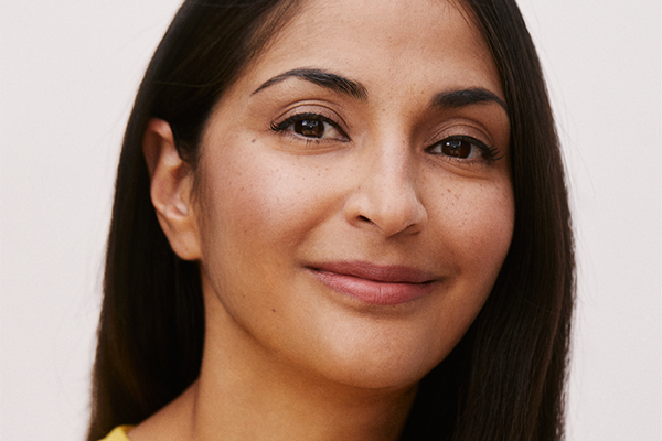 A woman with long dark hair smiles gently at the camera. She has brown eyes, clear skin, and wears a yellow top. The background is plain and light-colored.