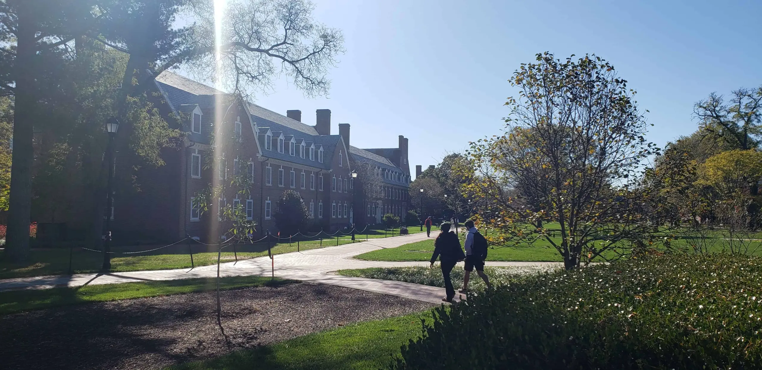 Two people walk along a sunlit path near a large brick campus building, surrounded by green lawns and trees on a clear day.