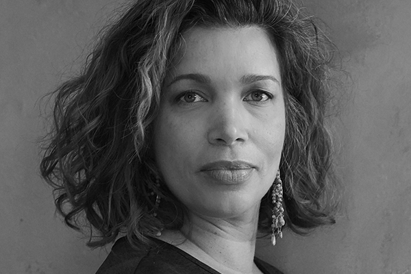 Black and white portrait of a woman with curly shoulder-length hair, looking directly at the camera. She is wearing earrings and a dark top, with a neutral expression and a plain background.