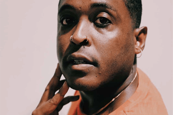 A person with short dark hair, wearing an orange shirt, gold necklace, nose ring, and small hoop earring, looks at the camera with one hand resting on their neck against a plain background.