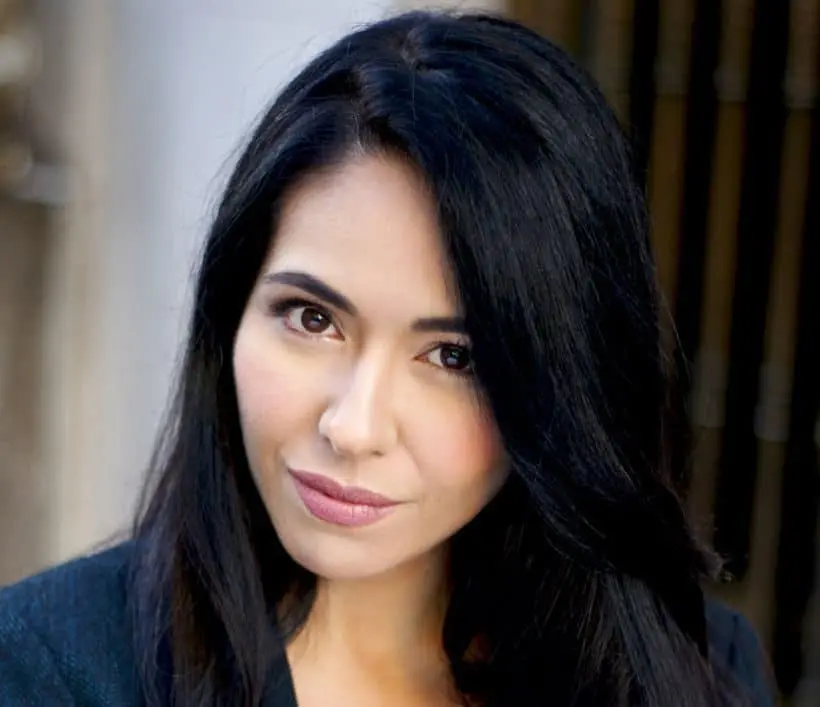 A woman with long, straight black hair and fair skin looks directly at the camera with a neutral expression, wearing a dark top. The background is blurred.
