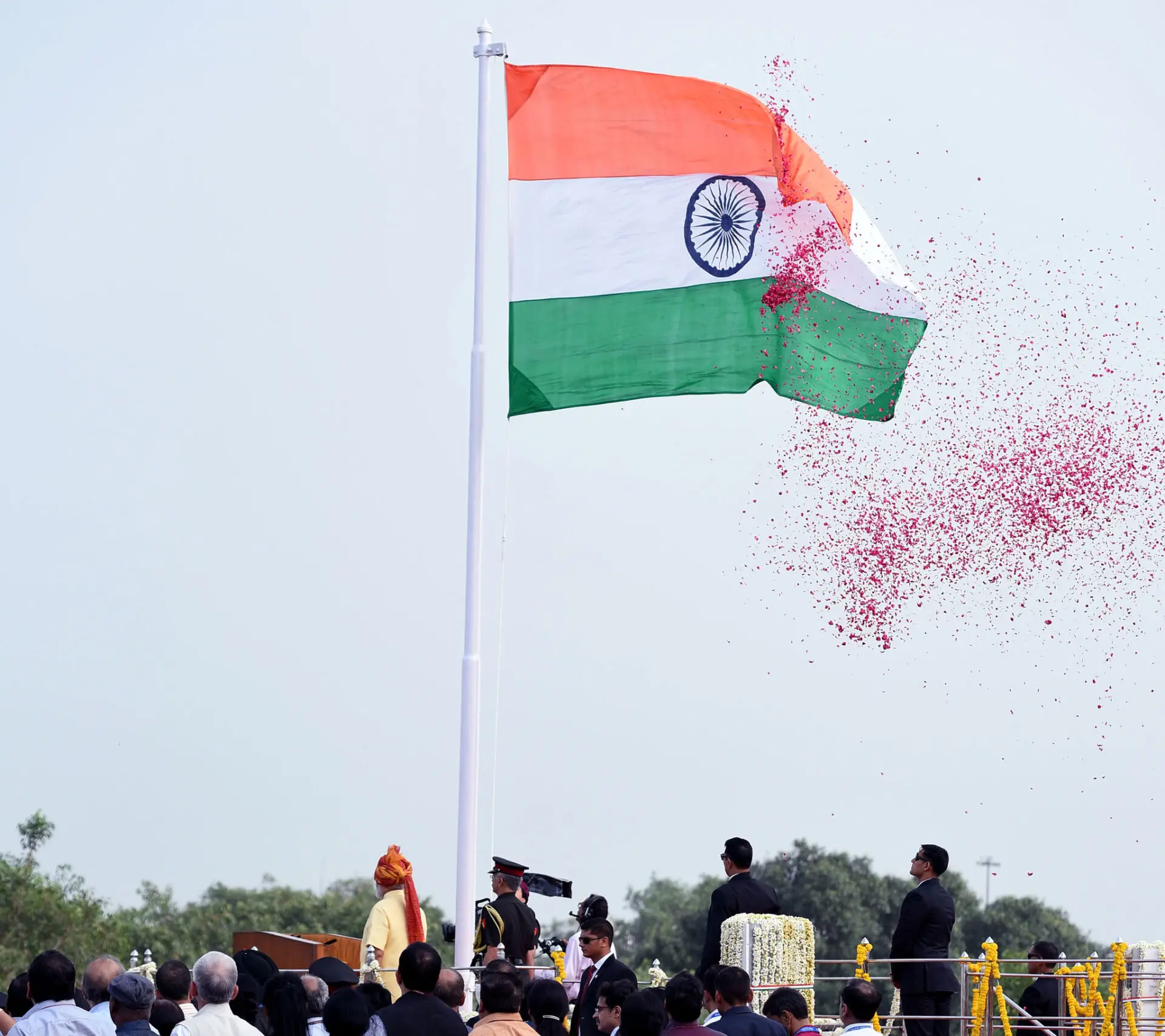 An Indian flag waves on a tall pole as rose petals are showered in the air. People, some in uniform, gather below, and a person in an orange turban stands at a podium during an India national event.