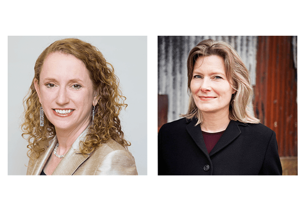 Two women facing the camera. The woman on the left has curly light brown hair and is wearing a beige blazer. The woman on the right has straight, light blonde hair and is wearing a black coat, with a corrugated metal wall in the background.
