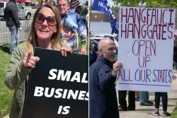Two people at a protest hold signs. One woman’s sign reads, SMALL BUSINESS IS, while a man’s sign says, HANG FAUCI HANG GATES OPEN UP ALL OUR STATES. Other people and flags are visible in the background.
