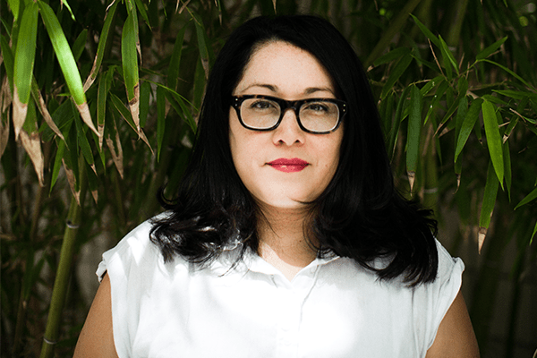 A woman with long dark hair and glasses, wearing a white shirt, stands in front of green bamboo leaves, looking directly at the camera with a neutral expression.
