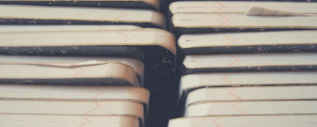 A close-up view of several stacked notebooks with worn edges and visible binding, reminiscent of prison writing, some with slightly curled pages, arranged in two uneven vertical piles.