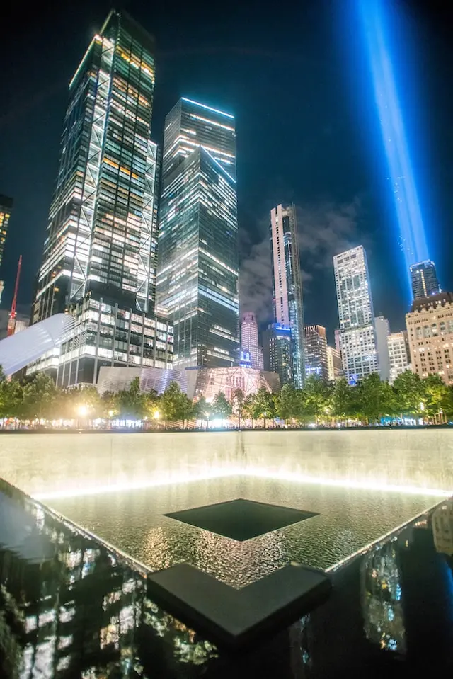 A glowing 9/11 Memorial reflecting pool at night with illuminated skyscrapers in the background and two blue beams of light shining into the sky in New York City.