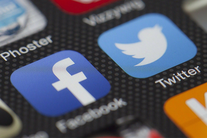 A close-up of smartphone screen showing app icons for Facebook and Twitter, with the blue Facebook f logo and the light blue Twitter bird logo clearly visible. Other app icons are partially seen around them.