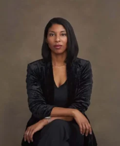 A woman with straight, shoulder-length black hair sits confidently with her hands crossed, embodying a strong sense of identity. She wears a black outfit and a velvet-textured blazer, posing against a plain brown background.