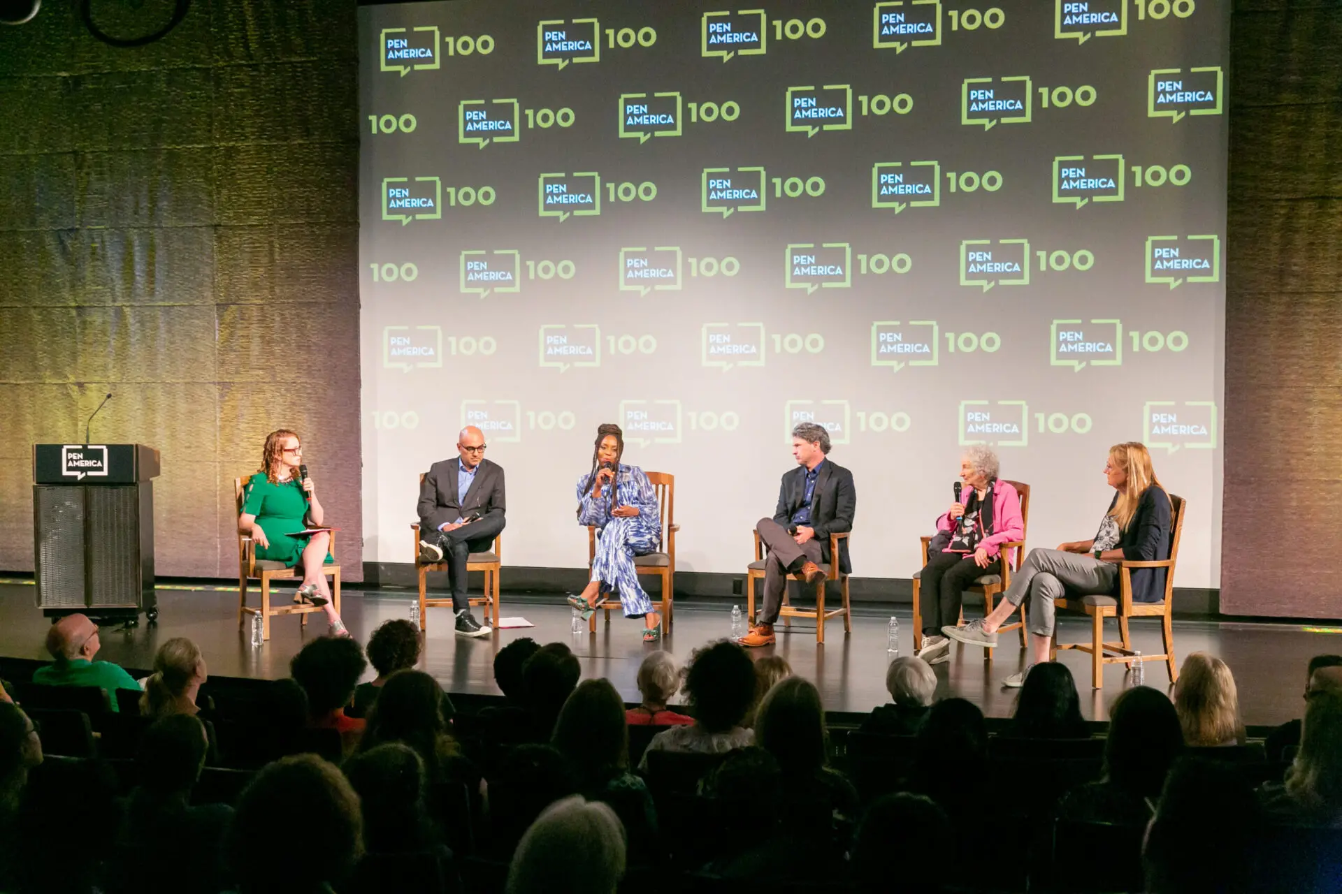 Six people sit on stage in front of an audience, participating in a panel discussion. A screen behind them displays the PEN America logo and the number 100 repeatedly.