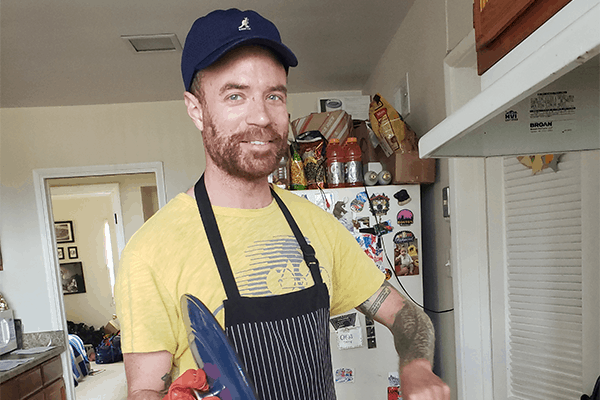 A smiling man with a beard, wearing a navy cap, yellow T-shirt, and striped apron, stands in a kitchen holding a blue plate. The refrigerator behind him is covered with magnets and photos.