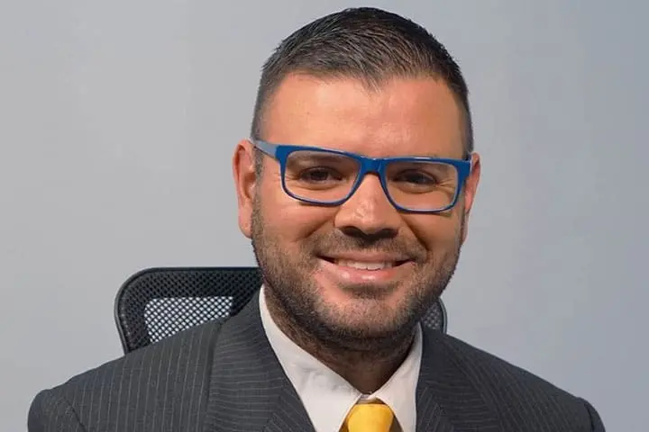 A man with short dark hair, a beard, and blue glasses is smiling. He is wearing a pinstripe suit, a white shirt, and a yellow tie, sitting in a black mesh office chair against a plain background.