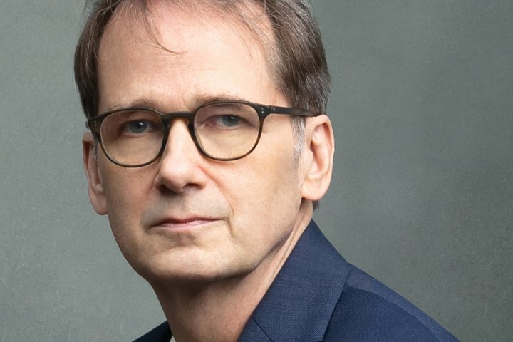 A middle-aged man with short brown hair and glasses, wearing a navy blue suit jacket, poses against a plain gray background.