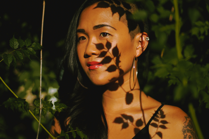 A person with long dark hair and hoop earrings stands among green leaves, with sunlight casting leaf-shaped shadows across their face and shoulder.