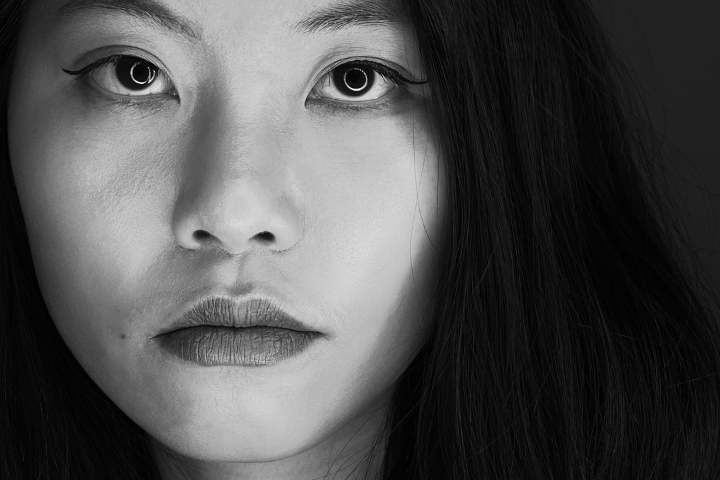 Black and white close-up of a woman’s face with long dark hair, neutral expression, and direct gaze. The image captures only the left side of her face, showing detailed skin texture and eye makeup.