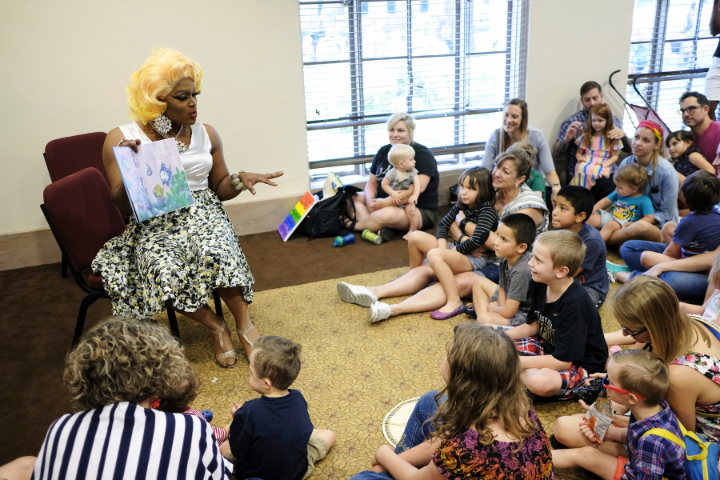A drag queen with blonde hair reads a picture book to a group of children and adults seated on the floor and chairs in a brightly lit room. A small rainbow flag is placed nearby.