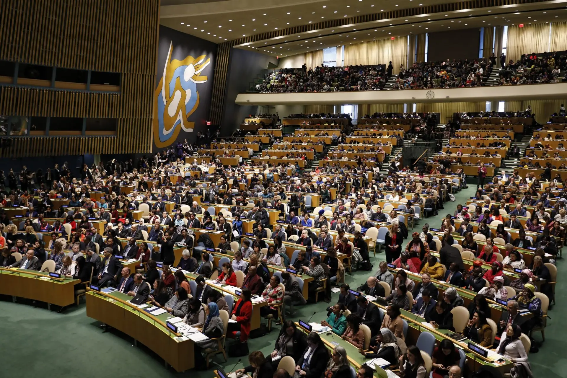 A large assembly hall filled with delegates seated at desks, facing a podium and stage. The room has a high ceiling, tiered seating, and a large abstract mural on the wall. The setting is formal and official.