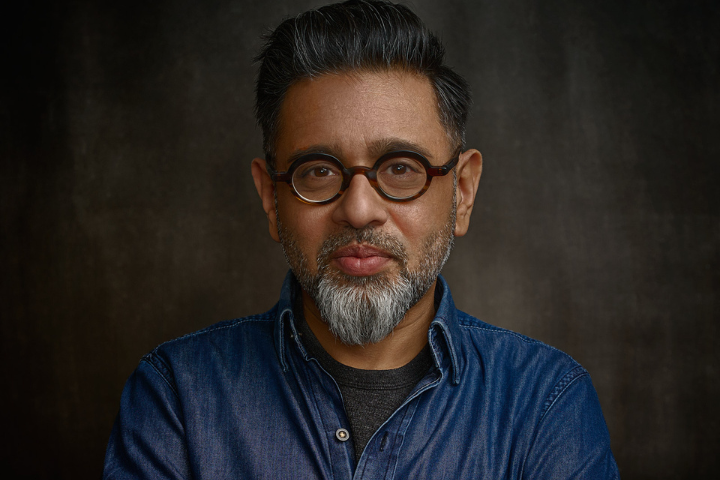 A middle-aged man with short dark hair, a gray beard, and mustache, wearing round glasses and a blue denim shirt, stands against a dark, textured background. He is looking directly at the camera and smiling slightly.