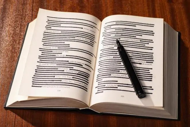 An open book lies on a wooden surface with most of the text on both pages blacked out by marker. A black marker pen rests across the right page.
