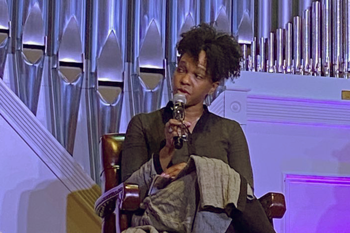 A woman with curly hair, dressed in black, sits in a chair holding a microphone and speaking on stage. Behind her are tall, shiny organ pipes and purple lighting.