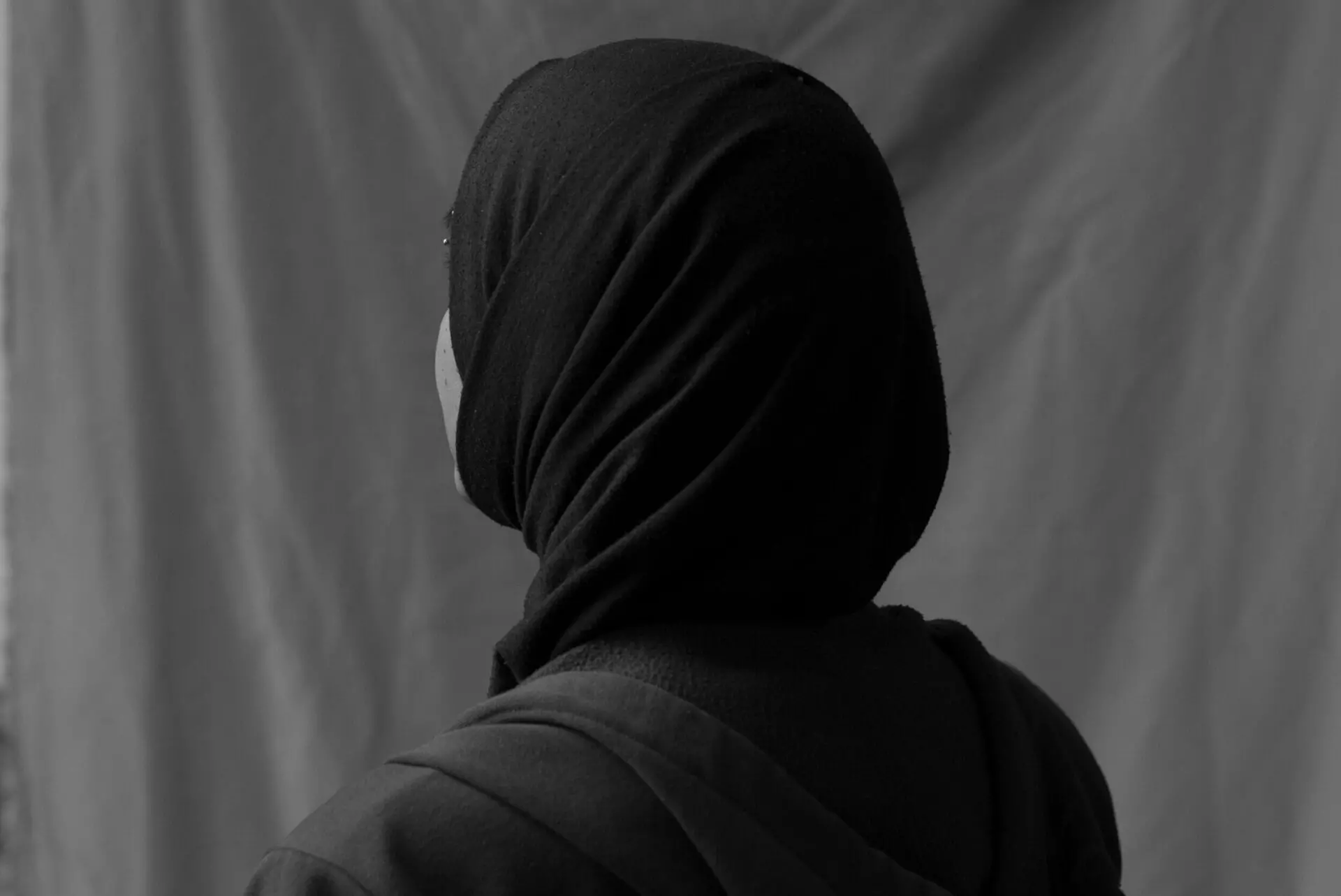 Black and white photo of a person wearing a dark hijab and cloak, seen from behind, facing a plain draped background.