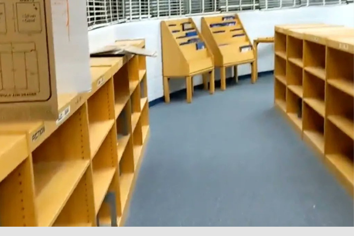 Empty wooden bookshelves and display stands are arranged in a room with blue carpet and white walls. Some boxes and cardboard are placed on top of the shelves.