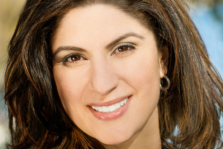 A woman with long brown hair, wearing hoop earrings, smiles warmly at the camera in a close-up outdoor portrait.