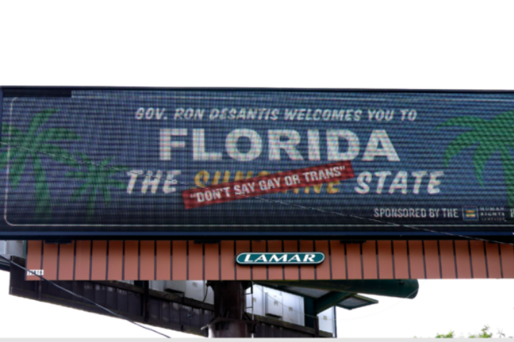 A billboard reads, Gov. Ron DeSantis welcomes you to Florida, The Sunshine State, with Dont Say Gay or Trans stamped in red text across it. Palm trees and sponsor logos are also visible.