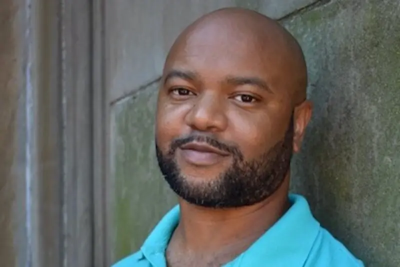 A man with a shaved head and trimmed beard wearing a light blue collared shirt stands against a textured stone wall, looking calmly at the camera.