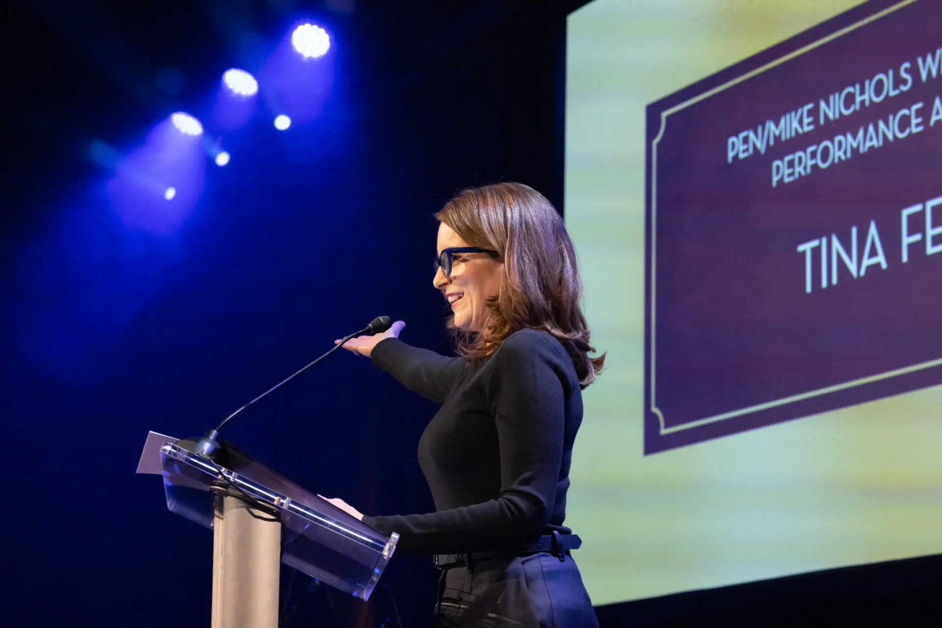 A woman in black glasses and a black outfit stands at a podium on stage, smiling and gesturing toward a screen that reads PEN/Mike Nichols Writing for Performance Award: Tina Fey. Blue stage lights shine above.