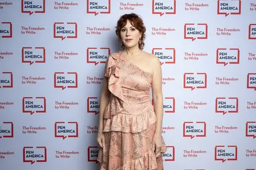 A woman in a pink, one-shoulder ruffled dress stands in front of a repeating PEN America The Freedom to Write backdrop at an event.