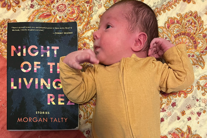 A baby in a yellow onesie lies on a patterned blanket next to the book Night of the Living Rez by Morgan Talty. The baby is looking to the side with arms bent upward.