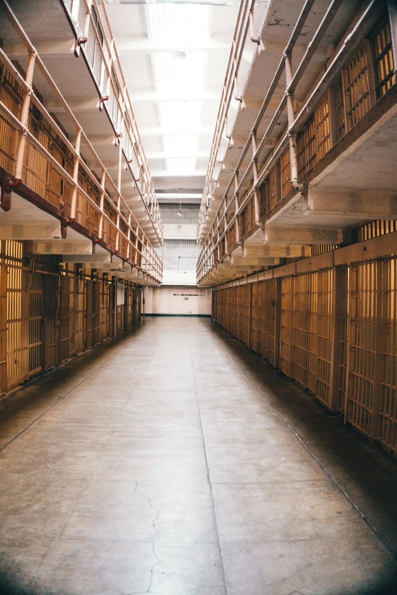 A long, empty corridor in a prison with rows of metal-barred cells on both sides and two levels, lit by overhead fluorescent lights.
