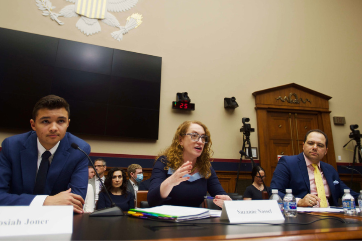 Three people sit at a panel table during a congressional hearing. The woman in the center speaks while the two men beside her listen. An audience and photographers are in the background. Nameplates and microphones are visible on the table.