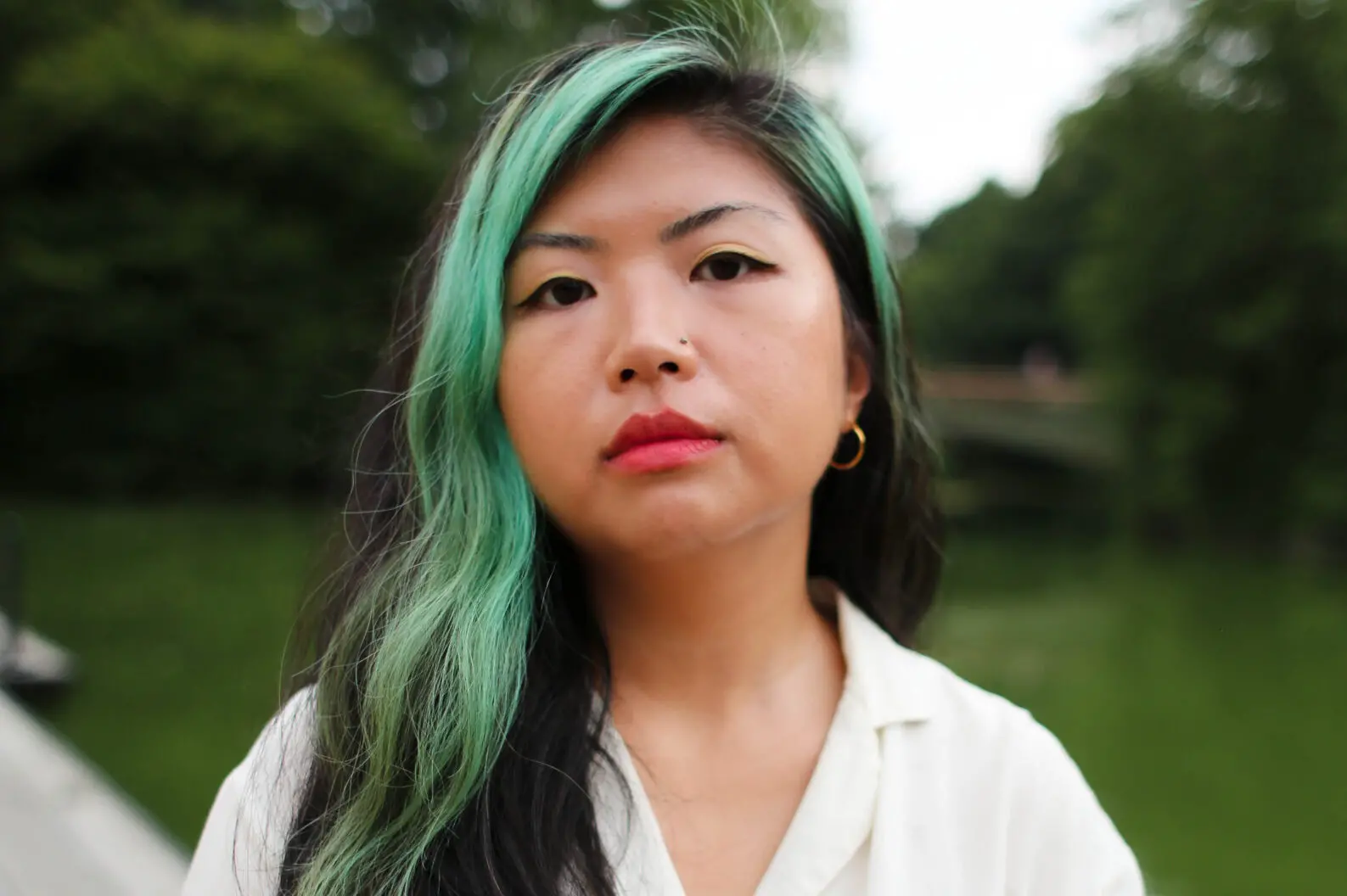 A woman with long, wavy hair dyed black and mint green stands outdoors, wearing a white shirt and gold hoop earring, looking directly at the camera with a neutral expression. Green trees and grass are in the background.