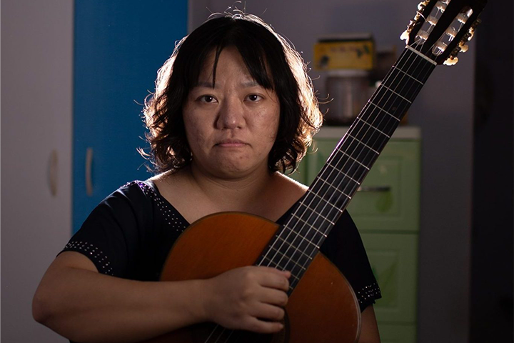A person with short, wavy hair holds a classical guitar and looks directly at the camera with a neutral expression. They are indoors, with a blue wardrobe and green drawers visible in the background.