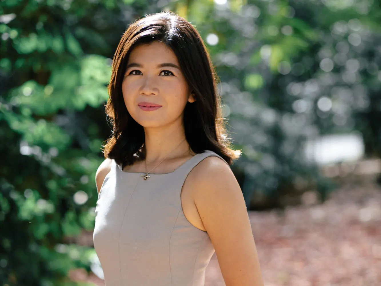 A woman with shoulder-length dark hair, wearing a sleeveless light-colored dress, stands outdoors with blurred green foliage and sunlight in the background. She looks slightly to the side with a calm expression.