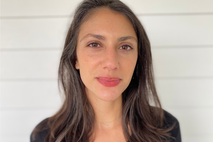 A woman with long brown hair and light skin stands in front of a white shiplap wall, looking directly at the camera with a neutral expression. She is wearing a black top.