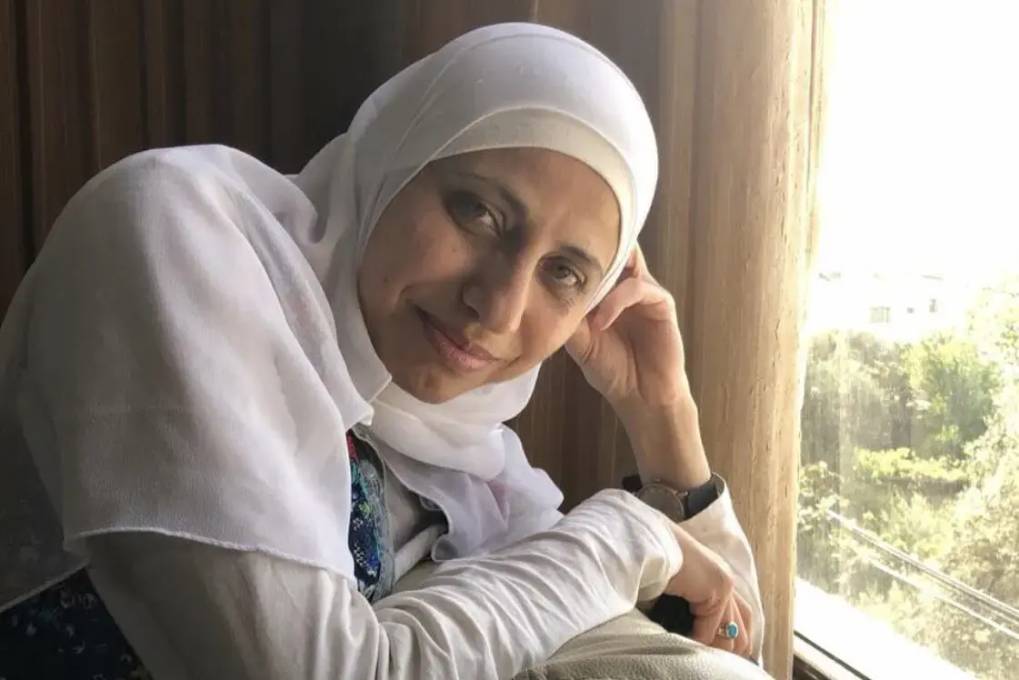 A woman wearing a white hijab and light-colored clothing leans by a window, resting her head on her hand and smiling slightly. Sunlight streams in, illuminating her face and the greenery outside.