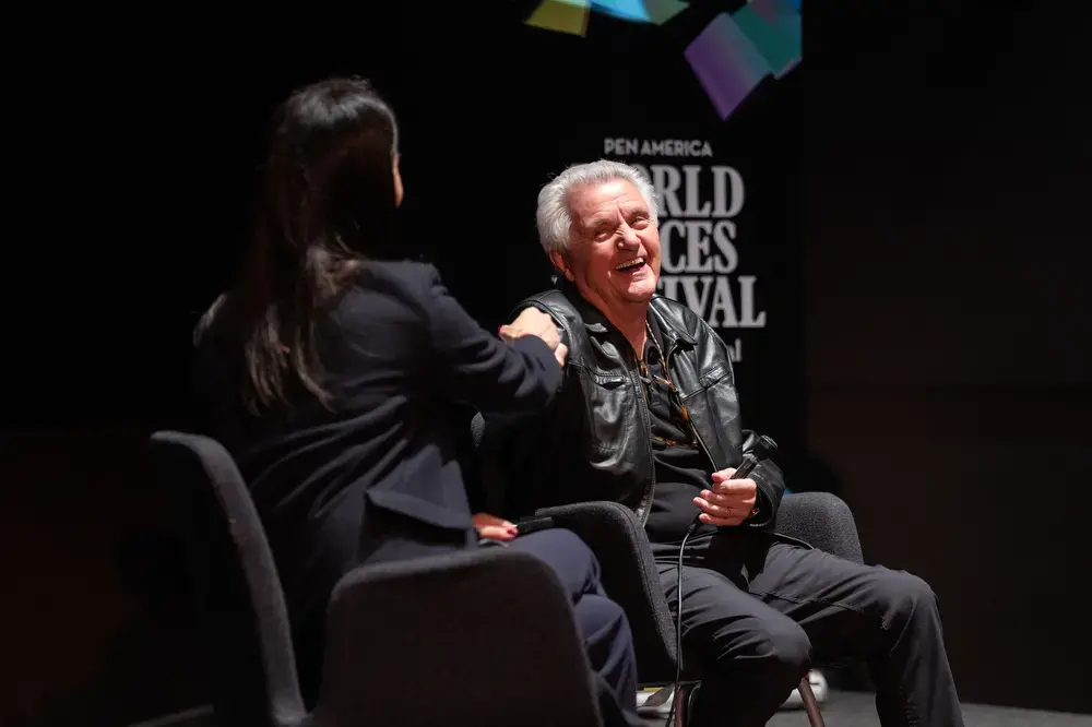 Two people sit on stage in conversation at the PEN America World Voices Festival. One, holding a microphone and wearing a leather jacket, smiles and laughs as the other gestures toward them.
