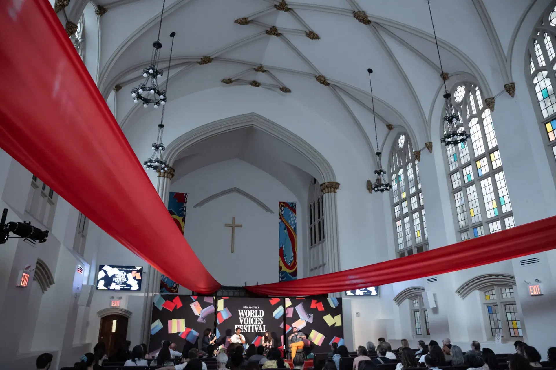 A large audience sits in a church-like hall with arched ceilings, red drapes, and tall windows, attending the World Voices Festival 2023 event on a stage decorated with colorful books and banners.