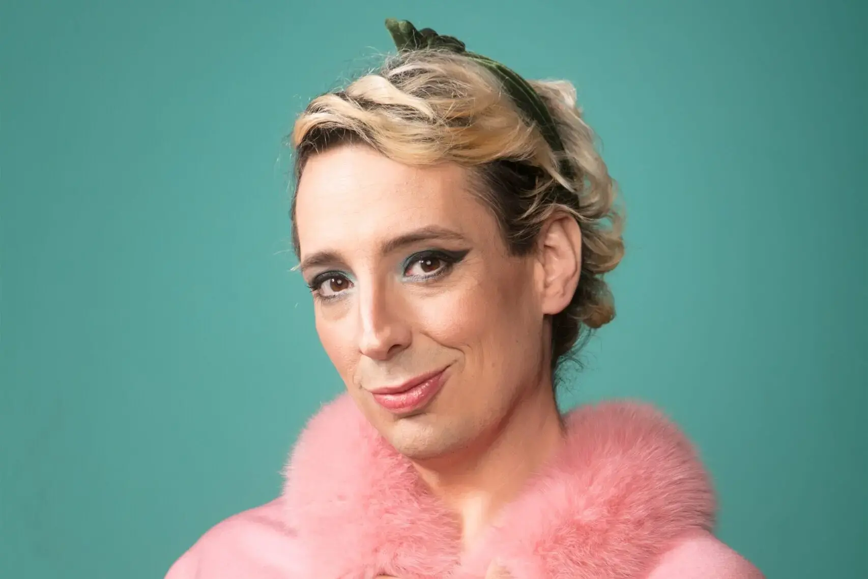 A person with wavy blond hair styled back, wearing winged eyeliner and a pink fur-collared coat, stands in front of a teal background, smiling softly at the camera.