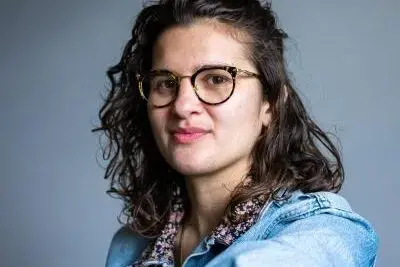 A person with wavy brown hair, wearing round eyeglasses, a floral shirt, and a light blue denim jacket, is looking at the camera, smiling slightly, with a neutral gray background.