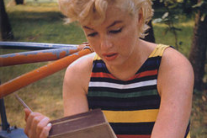 A person with short, curly blond hair wearing a colorful striped sleeveless top sits outdoors, looking down at an open book they are holding. Trees and grass are visible in the background.