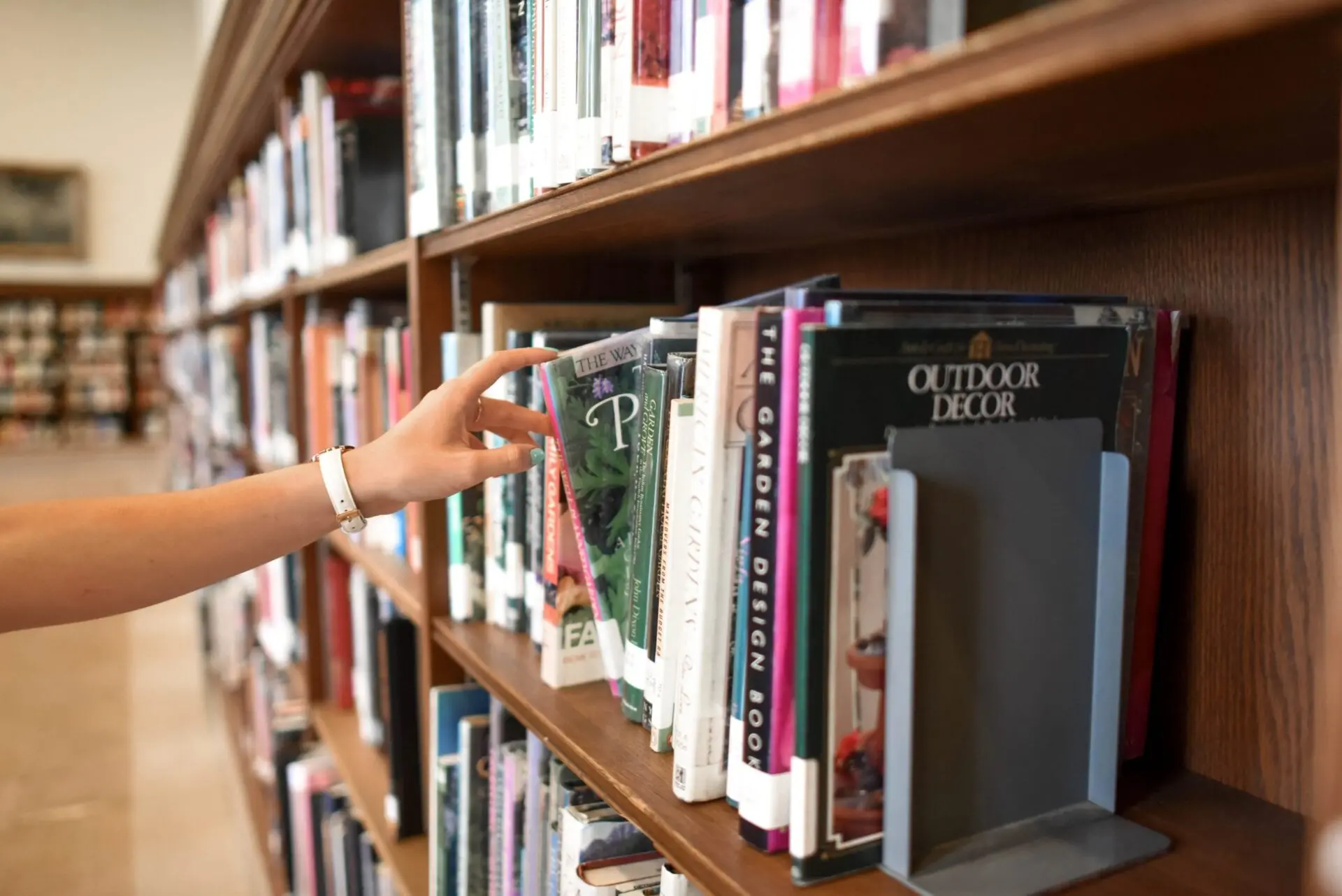 A person’s hand reaches for a book on a library shelf filled with various books, some with visible titles related to gardening and outdoor decor.